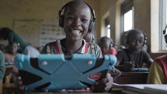 A smiling child wearing headphones and using a tablet in a classroom setting, showing excitement and engagement in digital learning.